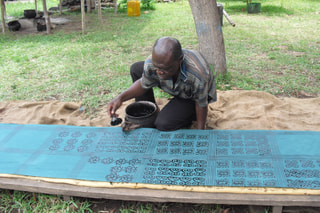 A craftsman kneeling to stamp Adinkra symbols onto cloth with a calabash stamp and dark dye