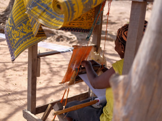 A weaver working at a narrow-strip loom, with finished Aso Oke cloth hanging above